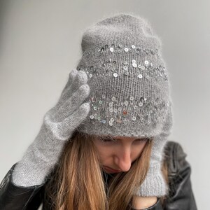 May include: A gray knit hat and matching gloves set. The hat is adorned with rows of silver sequins. The gloves are fingerless. The set is made of a soft, fuzzy material. The background is a solid light gray.