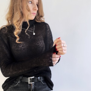 Pu&ograve; includere: Un maglione nero a maniche lunghe, semi-trasparente, con collo alto. Il maglione &egrave; abbinato a un paio di jeans grigio scuro con una cintura in pelle nera dotata di una fibbia rettangolare color argento. Una collana e un anello in argento con motivo a quadrifoglio.
