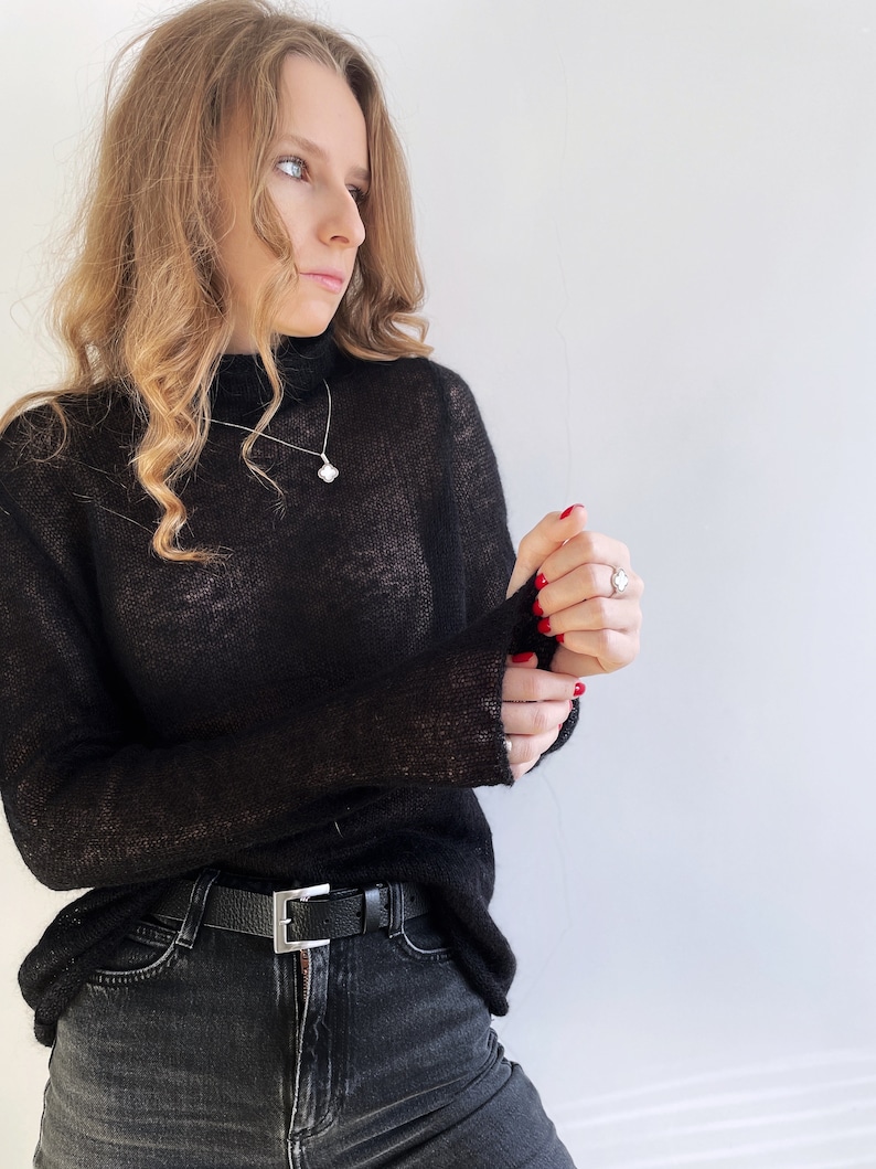 Pu&ograve; includere: Un maglione nero a maniche lunghe, semi-trasparente, con collo alto. Il maglione &egrave; abbinato a un paio di jeans grigio scuro con una cintura in pelle nera dotata di una fibbia rettangolare color argento. Una collana e un anello in argento con motivo a quadrifoglio.