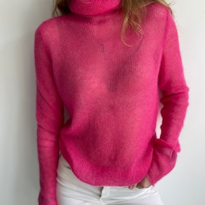 Può includere: Un maglione rosa trasparente a collo alto con una vestibilità ampia. Il maglione è realizzato in un tessuto leggero e delicato.
