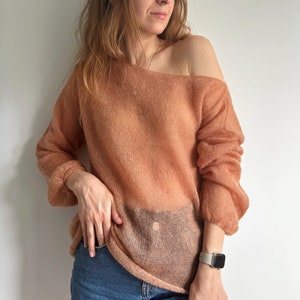 Könnte beinhalten: Ein hellpfirsichfarbener, offener Pullover mit lockerer Passform und langen Ärmeln. Der Pullover ist aus einem weichen, leichten Material gefertigt und hat eine leicht durchscheinende Optik.