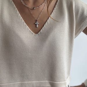 Puede incluir: Una camiseta de punto beige con cuello en V combinada con un collar en capas con un colgante de cruz. La camiseta es de color neutro y la joyería añade un toque delicado. El estilo general es sencillo y elegante.
