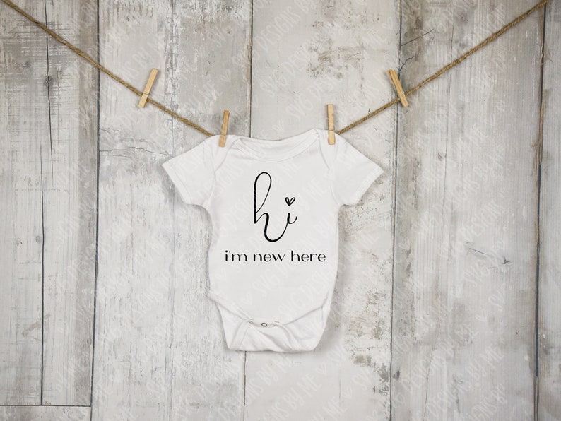 May include: White baby onesie with the text "hi i'm new here" in black script. The onesie is hanging from a clothesline with wooden clothespins against a weathered, white wooden background.