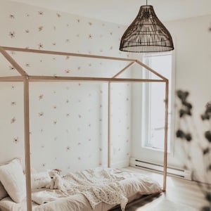 May include: A wooden canopy bed frame with a white floral patterned wallpaper in the background. The bed is made with a white and beige bedding set and a stuffed animal is on the bed. A woven pendant light hangs above the bed.