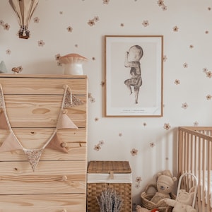 May include: A light wood dresser with a floral garland, a wicker basket with lavender, and a white ceramic jug. A framed print of a child in a pose hangs on the wall above the dresser. The wall is decorated with small brown flower decals.