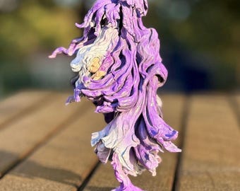 May include: A handcrafted sculpture of a figure with flowing, layered forms in shades of purple and white. The artwork has a textured appearance and stands on a wooden surface, with a blurred background.