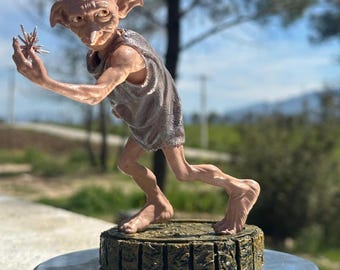 May include: A detailed figurine of a character with large ears, a long nose, and wearing a gray tunic. The figure is holding a small, star-shaped object in its outstretched hand. It stands on a textured, rectangular base.