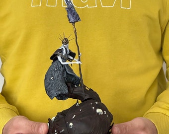 May include: A detailed figurine of a spectral figure holding a staff with a tattered banner. The figure is dressed in dark robes and a crown, standing atop a dark, textured base adorned with skulls. The figurine is held against a yellow hoodie.