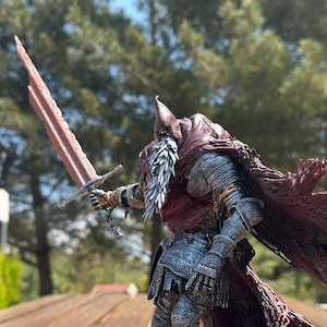 Op de afbeelding: Een gedetailleerd actiefiguur van een ridder in zilveren harnas met een donkerrode, gerafelde mantel. Het figuur heeft een puntige kap, een lange witte baard en houdt een groot, gekarteld, roodbruin zwaard uitgestrekt in de rechterhand.