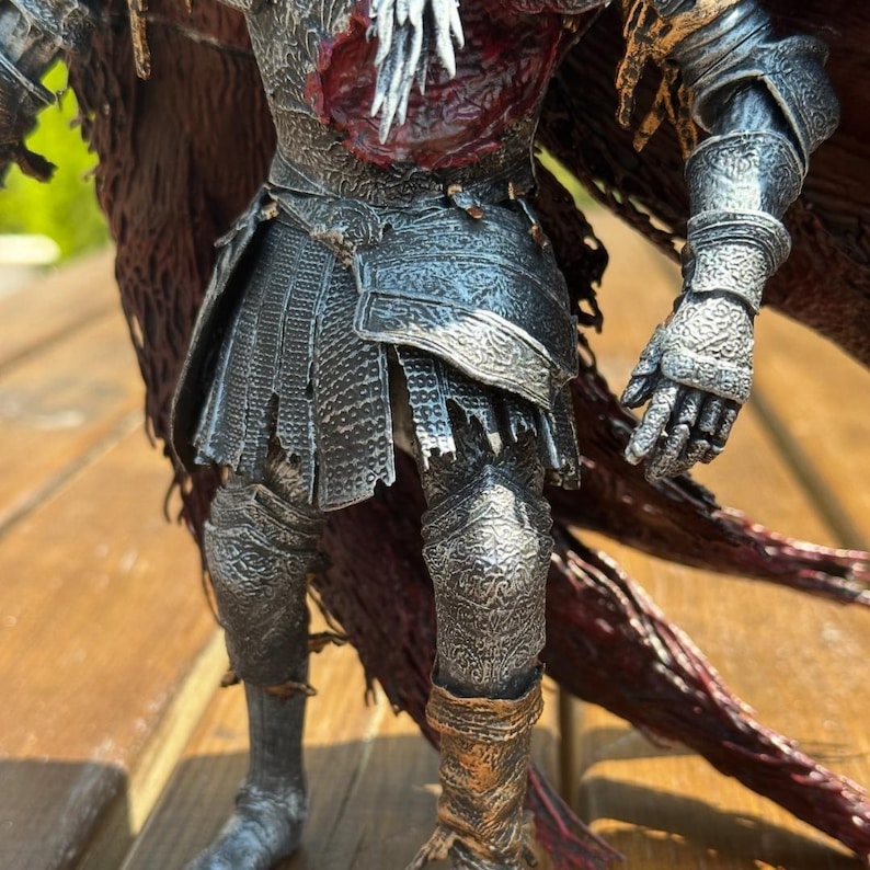 Op de afbeelding: Een gedetailleerd actiefiguur van een krijger in ingewikkeld, verweerd zilverkleurig en donkergrijs harnas. Het figuur heeft een gehavende, donkerrode cape, een borststuk met een textuurrijke rode wond en gedetailleerde gravures op de handschoenen.