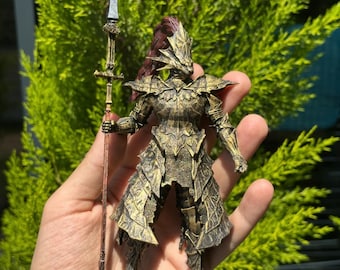 May include: A detailed figurine of a knight in ornate armor, holding a spear. The armor is a mix of gold and bronze tones, with intricate patterns. The knight has a helmet with a red plume. The figurine is held in a hand against a backdrop of green foliage.