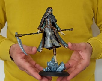 May include: A detailed figurine of a robed figure with a scythe, standing on a blue crystal base. The figure wears a dark cloak and a long dress with gold and teal accents. The scythe is held in both hands.