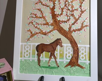 May include: A framed artwork featuring a brown horse under a tree with pink and green leaves. The tree has a textured brown trunk. A white fence and a textured green ground are in the foreground. The artwork is in a white frame.