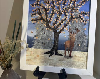 May include: Framed artwork depicting a deer standing under a tree with autumn-colored leaves. The background features a full moon and snow-covered trees. The artwork is in a white frame and displayed on a black easel.