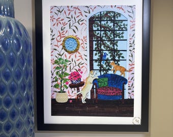 May include: A framed art print featuring a room with patterned wallpaper, a large arched window with a blue bird, a yellow dog standing by a blue armchair, an orange cat on the chair, a potted plant, and a small round gold-framed wall mirror.