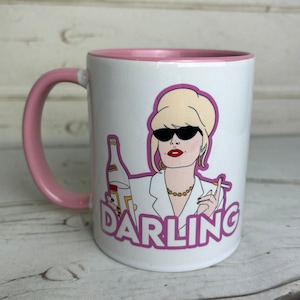 May include: White ceramic mug with a pink rim and handle. The mug features a cartoon illustration of a woman with blonde hair wearing sunglasses and a white shirt. She is holding a bottle of alcohol and a cigarette. The text "DARLING" is printed in purple below the illustration.
