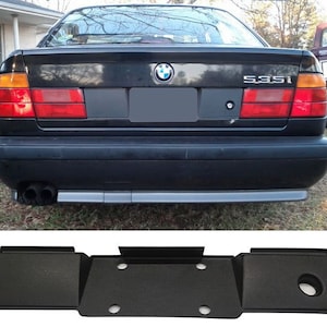 Könnte beinhalten: Schwarzer Kunststoff-Heckstoßfänger-Ersatz für einen BMW 535i. Der Stoßfänger hat eine glatte, flache Oberfläche mit einem rechteckigen Ausschnitt für ein Nummernschild. Es befinden sich vier runde Löcher am Stoßfänger.