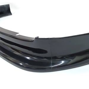 Puede incluir: Un aler&oacute;n de labio de parachoques de coche de pl&aacute;stico negro. El aler&oacute;n es curvo y tiene un acabado liso y brillante.