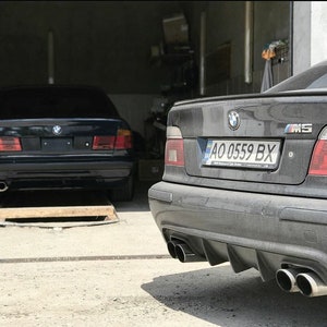May include: Two black BMW saloons parked in a garage. The car in the foreground has a number plate that reads "AO 0559 BX" and an "M5" badge on the boot. Both cars have dual exhausts.
