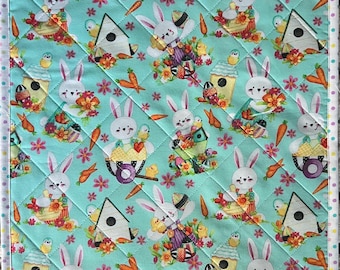 May include: A quilted square featuring a repeating pattern of Easter bunnies, carrots, birdhouses, and flowers on a light blue background. The design includes a white border with small, colorful polka dots.