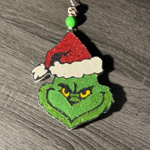 May include: A glittery green Grinch face ornament with a red Santa hat and white trim. The ornament has yellow eyes and a white string for hanging. A green bead and a patterned bead are attached to the string.