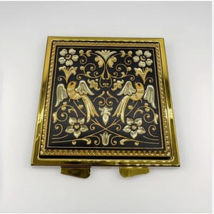 May include: A square, decorative metal box with a black background and gold accents. The box features a detailed design of two birds, flowers, and ornate scrollwork. The frame is gold-colored, and the box has small feet.