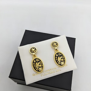 Puede incluir: Pendientes dorados con detalles de esmalte negro. Cada pendiente presenta un perno circular con un diseño de pájaro y un colgante ovalado colgante con un motivo de pájaro y floral. Los pendientes se exhiben sobre un cojín blanco dentro de una caja negra.