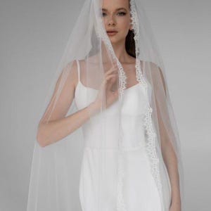 Puede incluir: Un velo de novia blanco con delicados bordes de encaje, sobre un vestido de novia blanco. El velo es transparente, creando una apariencia suave y etérea. El vestido tiene un diseño simple y elegante, perfecto para una ceremonia de boda.