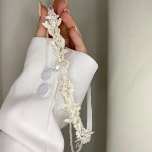May include: A white bridal headband featuring delicate floral embellishments, pearls, and crystal beads. The headband is decorated with a chain detail and a white ribbon.