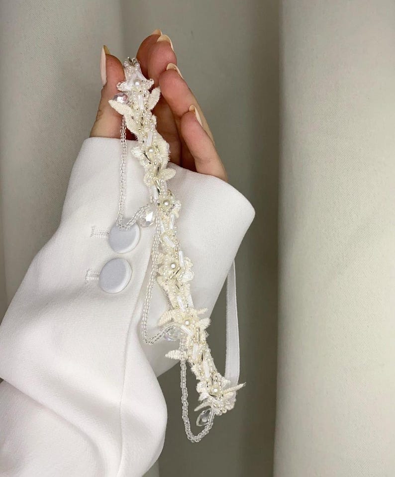 May include: A white bridal headband featuring delicate floral embellishments, pearls, and crystal beads. The headband is decorated with a chain detail and a white ribbon.