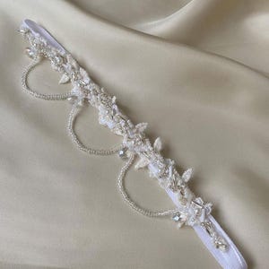 May include: White bridal garter featuring delicate beaded lace and pearl embellishments. The garter has a white elastic band and a decorative beaded design.