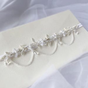 May include: A delicate white and silver garter with a floral design and dangling chains of beads. The garter is on a white fabric background.