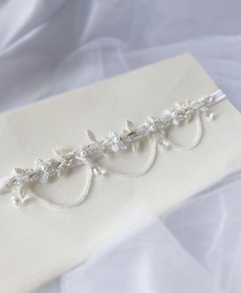 May include: A delicate white and silver garter with a floral design and dangling chains of beads. The garter is on a white fabric background.