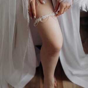 May include: A white bridal garter with delicate lace and pearl embellishments. The garter is worn on the leg of a person.