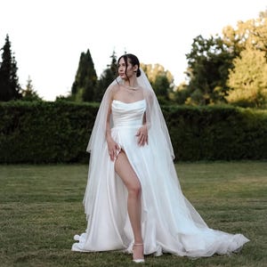 May include: A white strapless wedding dress with a high leg slit, complemented by a sheer veil. The dress has a flowing skirt and is worn with white heels. The image is captured outdoors on a grassy lawn.