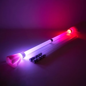 May include: A glowing, fiber optic light wand with a pink and purple illuminated brush-like end. The wand is white with black accents and is accompanied by two smaller, decorative wands. The background is a gradient of purple and red.