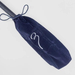 May include: A dark blue velvet protective cover for a cricket bat, featuring a drawstring closure at the top and a white embroidered letter M on the front. The handle of the cricket bat is visible extending from the top of the fabric pouch.