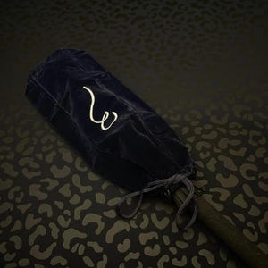 May include: A navy blue velvet umbrella case with a white embroidered "w" logo. The case is closed with a drawstring and is partially covering a dark-colored umbrella handle. The background has a dark leopard print.