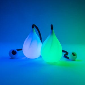 May include: Two teardrop-shaped LED lamps with smooth, translucent white surfaces that glow in blue and green light. Each lamp features a black braided cord attached to the top, with a small white spherical bead at the end of each cord.