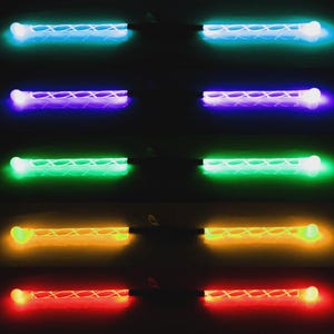 Puede incluir: Una colección de barras de luz LED brillantes en varios colores, incluyendo azul, morado, verde, naranja y rojo. Cada barra tiene un extremo blanco y un patrón entrecruzado a lo largo del tubo iluminado. El fondo es oscuro.