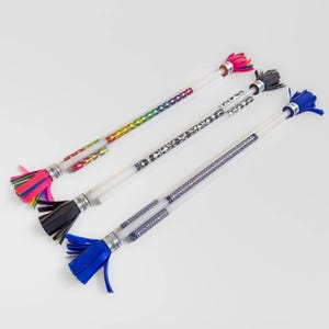 Peut inclure: Quatre bâtons de jonglage colorés avec des tiges en plastique transparent et des motifs. Chaque bâton a une touffe de couleur différente à l'extrémité : rose, noir, bleu et un arc-en-ciel de couleurs. Les bâtons sont disposés en diagonale sur une surface blanche.