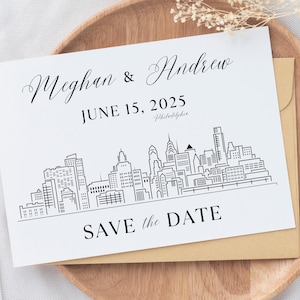 May include: A white save the date card with black text. The text reads "Meghan & Andrew" "June 15, 2025" "Philadelphia" and "Save the Date". The card features a black line drawing of the Philadelphia skyline.
