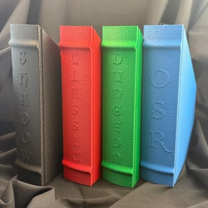 May include: Four colorful book-shaped storage boxes. The boxes are in shades of gray, red, green, and blue. Each box has embossed text on the spine: "SHADOW", "CHESSIS", "DUNGEON", and "OSR".