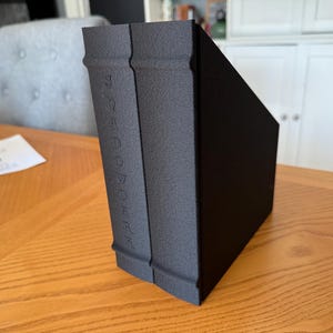 May include: A black book-shaped storage box with the word "BOOKARK" embossed vertically on the spine. The box is made of a textured material and is angled on a wooden table. The background includes a gray chair and a white cabinet.