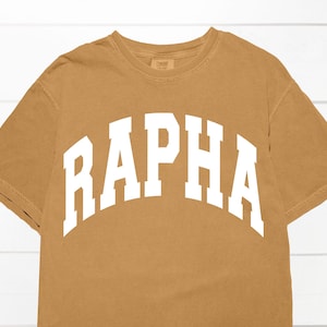 May include: A brown t-shirt with a white "RAPHA" graphic printed on the front.