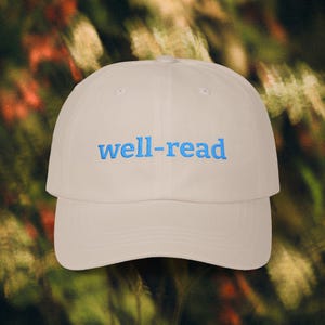 May include: A cream-coloured baseball cap with a curved peak. The words "well-read" are embroidered in blue across the front. The cap is made of a soft material and has a classic six-panel design.