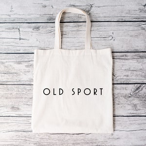 K&ouml;nnte beinhalten: Eine wei&szlig;e Canvas-Tragetasche mit schwarzen Henkeln und dem Text "OLD SPORT" in Schwarz auf der Vorderseite.