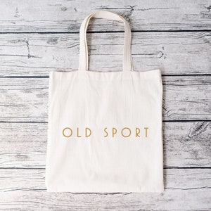 K&ouml;nnte beinhalten: Eine wei&szlig;e Canvas-Tragetasche mit dem Aufdruck "OLD SPORT" in Gold auf der Vorderseite.