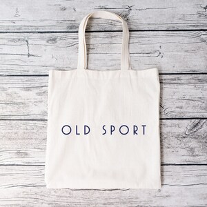 K&ouml;nnte beinhalten: Eine wei&szlig;e Einkaufstasche mit dem Text "OLD SPORT" in Blau auf der Vorderseite. Die Tasche liegt auf einer wei&szlig;en Holzoberfl&auml;che.