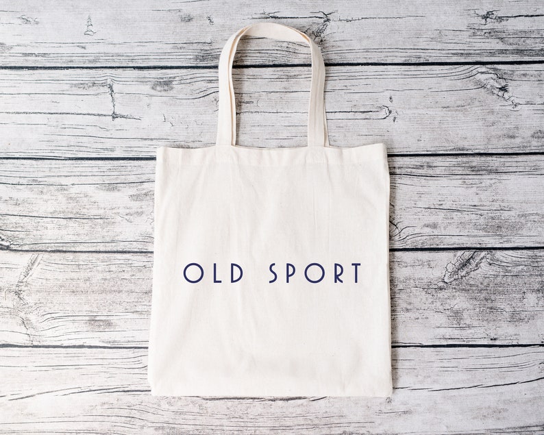 K&ouml;nnte beinhalten: Eine wei&szlig;e Einkaufstasche mit dem Text "OLD SPORT" in Blau auf der Vorderseite. Die Tasche liegt auf einer wei&szlig;en Holzoberfl&auml;che.
